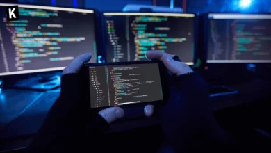 Hackeur examine du code sur un téléphone avec des écrans d'ordi en arrière-plan